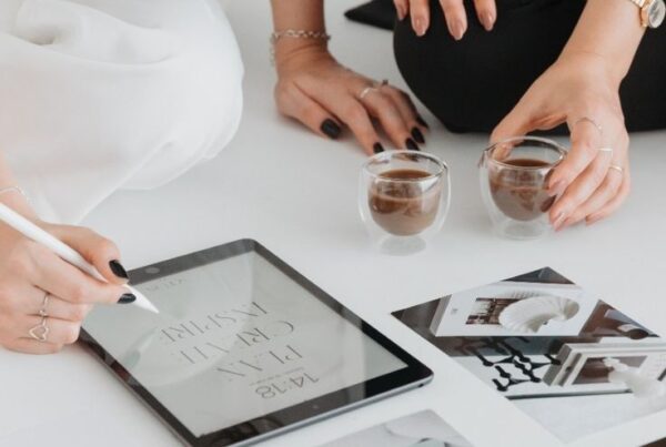 Two women collaborating on custom logo design in Newcastle, drawing on an iPad with branding images spread across a table.
