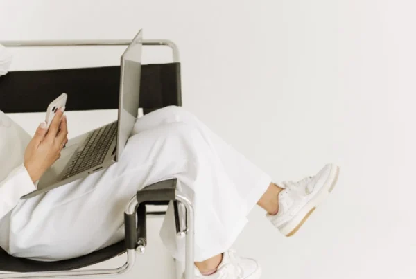 Woman relaxing with legs over the side of a chair while working on a laptop and holding an iPhone, representing modern creative work and branding strategy.