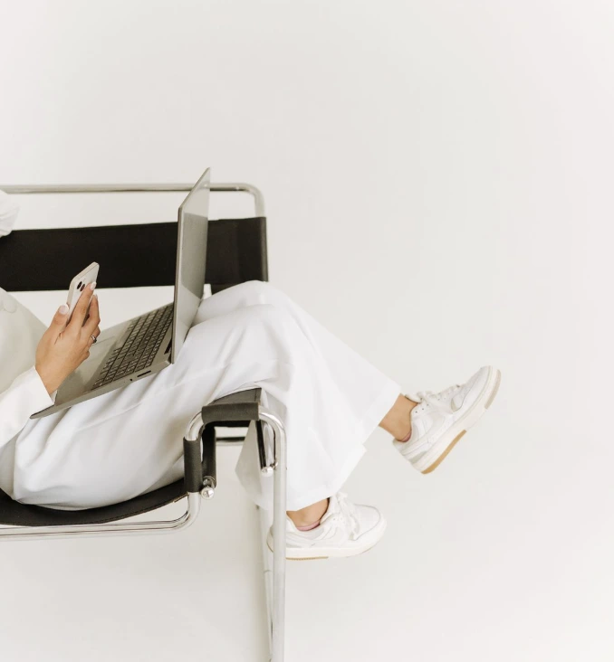 Woman relaxing with legs over the side of a chair while working on a laptop and holding an iPhone, representing modern creative work and branding strategy.